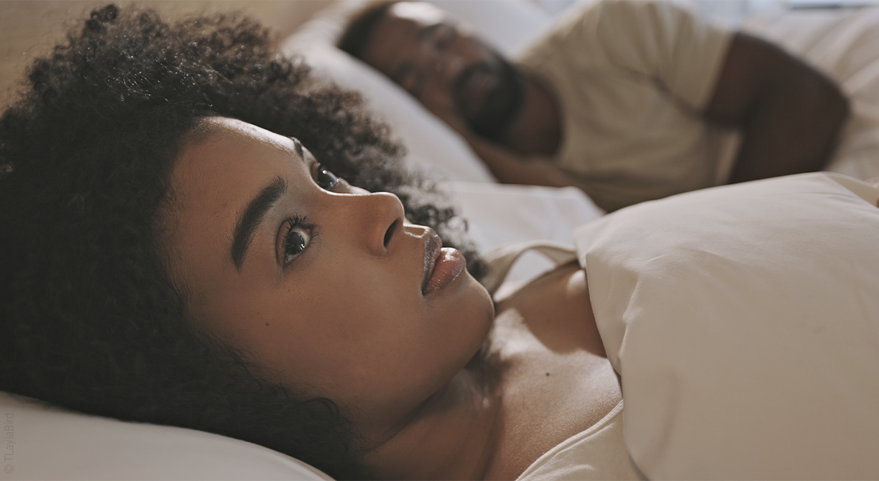 Titelbild Einschlafen in 60 Sekunden: Eine Person liegt zugedeckt mit geöffneten Augen im Bett, neben ihr liegt eine schlafende Person.