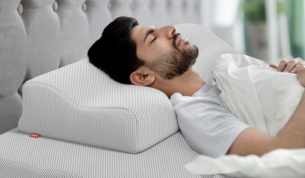 Foto: Eine Person liegt mit geschlossenen Augen auf dem Rücken im Bett, ihr Kopf ist auf dem BODYGUARD® Nackenstützkissen abgelegt