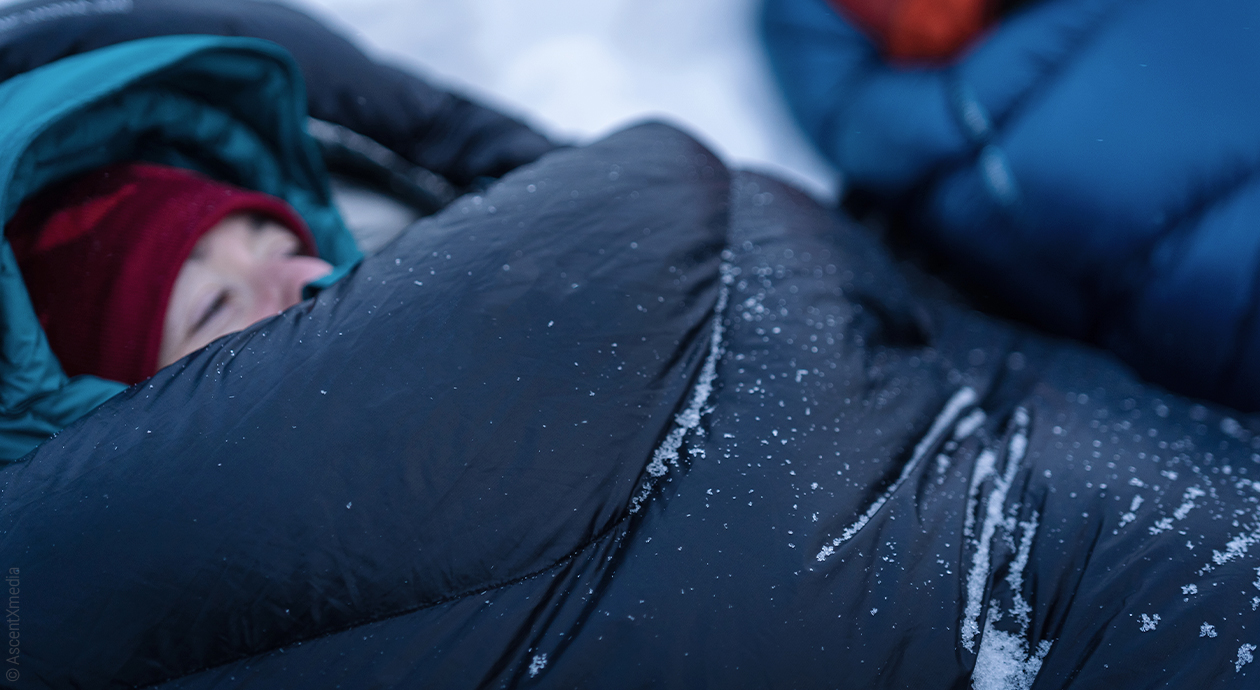 Titelfoto Schlafsack: Eine Person mit Mütze auf dem Kopf liegt in einem Schlafsack, auf dem Schneeflocken liegen.