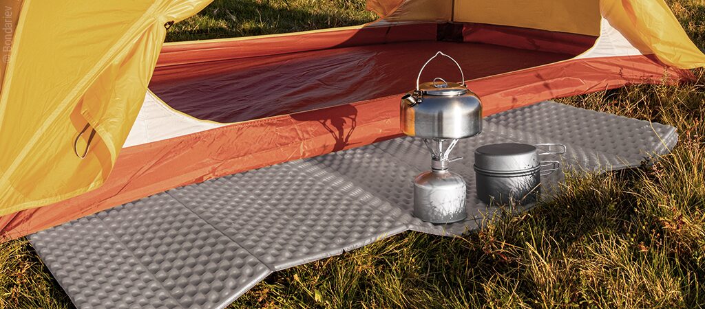 Foto: Eine faltbare Schaumstoff-Isomatte liegt vor einem geöffneten Zelt. Auf der Matte stehen ein Gaskocher mit Topf und daneben Camping-Geschirr.