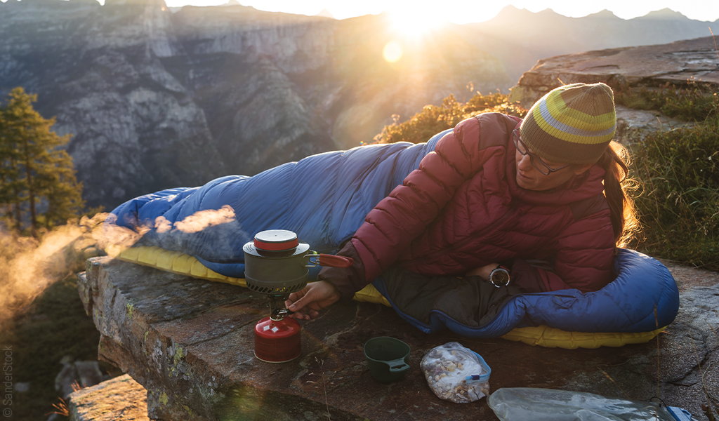 Foto: Eine Person in Winterkleidung liegt auf einer Isomatte auf einem Felsvorsprung. Neben ihr steht ein Gaskocher mit Topf. Im Hintergrund geht die Sonne hinter den Bergen auf.