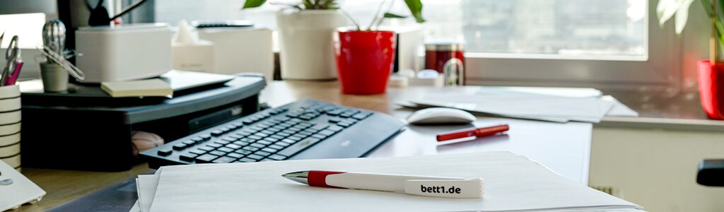 Foto: Ein Büro-Schreibtisch mit Maus und Tastatur. Daneben Notizzettel, darauf ein bett1 Kugelschreiber.