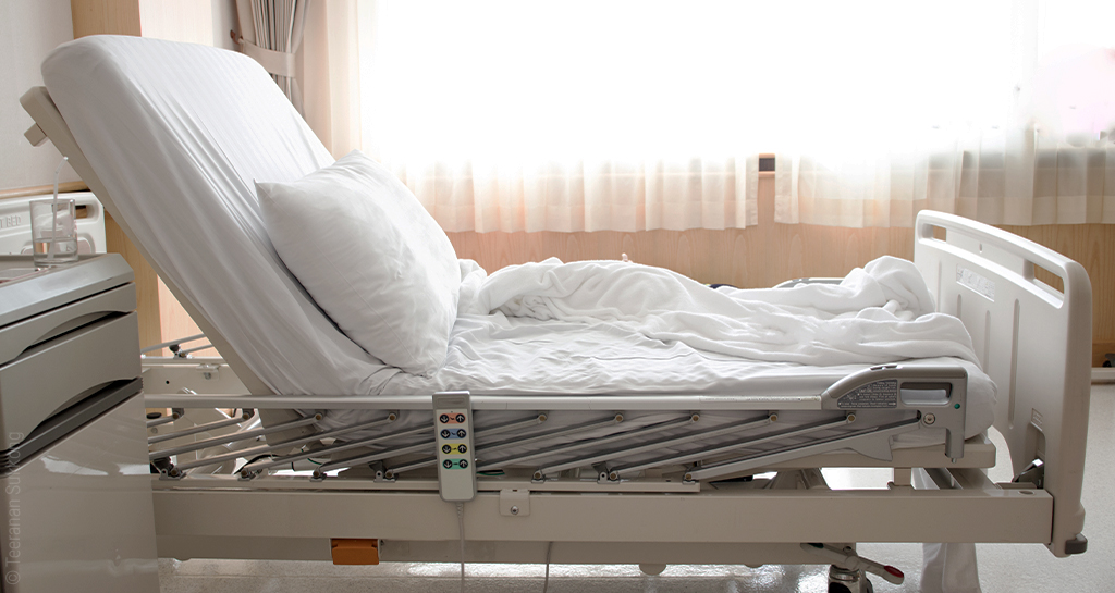 Foto: Ein elektrisch verstellbares Pflegebett mit hochgestelltem Kopfteil, daneben ein Krankenhaus-typischer Nachttisch.