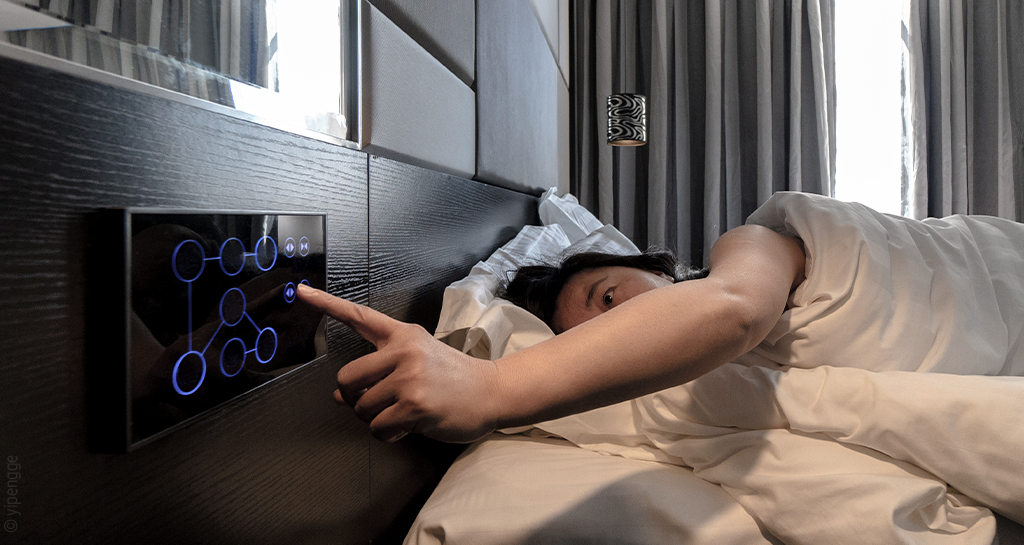 Foto: Eine Person liegt im Bett und tippt mit dem Finger auf ein Tablet, das im Kopfteil des Bettes eingebaut ist.