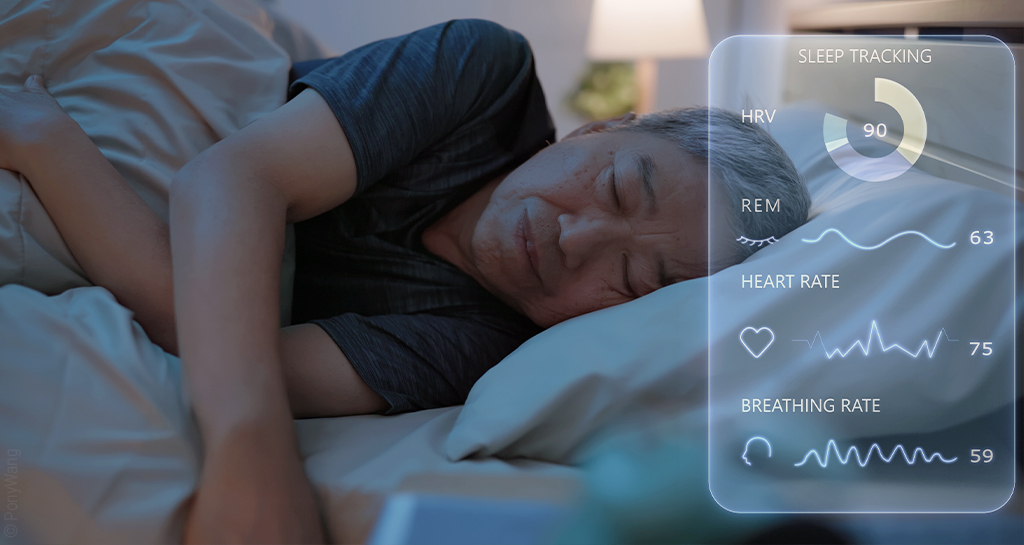 Foto: Eine Person liegt schlafend im Bett. Im Vordergrund ist ein transparentes, digitales Display, das Schlafdaten wie Herz-Frequenz und REM-Phase anzeigt. 