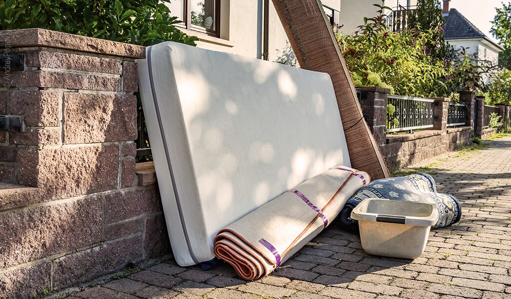 Foto: Eine schmutzige Matratze lehnt in einem Wohngebiet an einem Gartenzaun. Davor liegen zwei zusammengerollte Teppiche und weiteres Sperrgut.