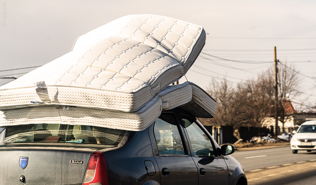 Foto: Zwei Matratzen sind auf einem Autodach festgeschnallt. Das Auto fährt durch eine Wohnsiedlung. 