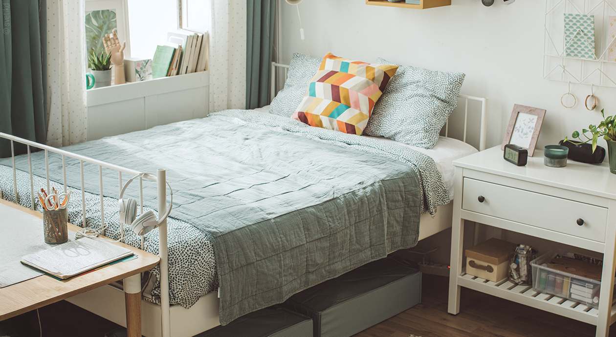 Foto: Auf einem ordentlich gemachten Einzelbett liegt eine Tagesdecke. Am Fußende steht ein Schreibtisch, auf dem Mal-Utensilien stehen. Dahinter eine Fensterbank, auf der Bücher stehen. Neben dem Bett ein Nachttisch.