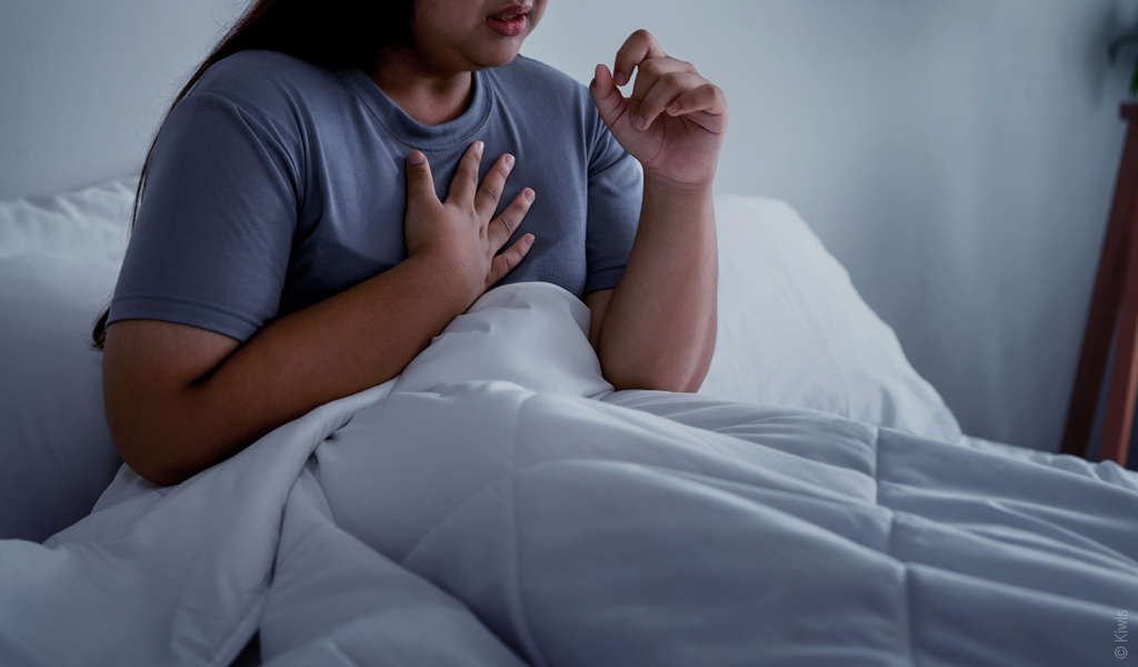 Eine Person sitzt aufrecht im Bett und hält sich eine Hand an die Brust.