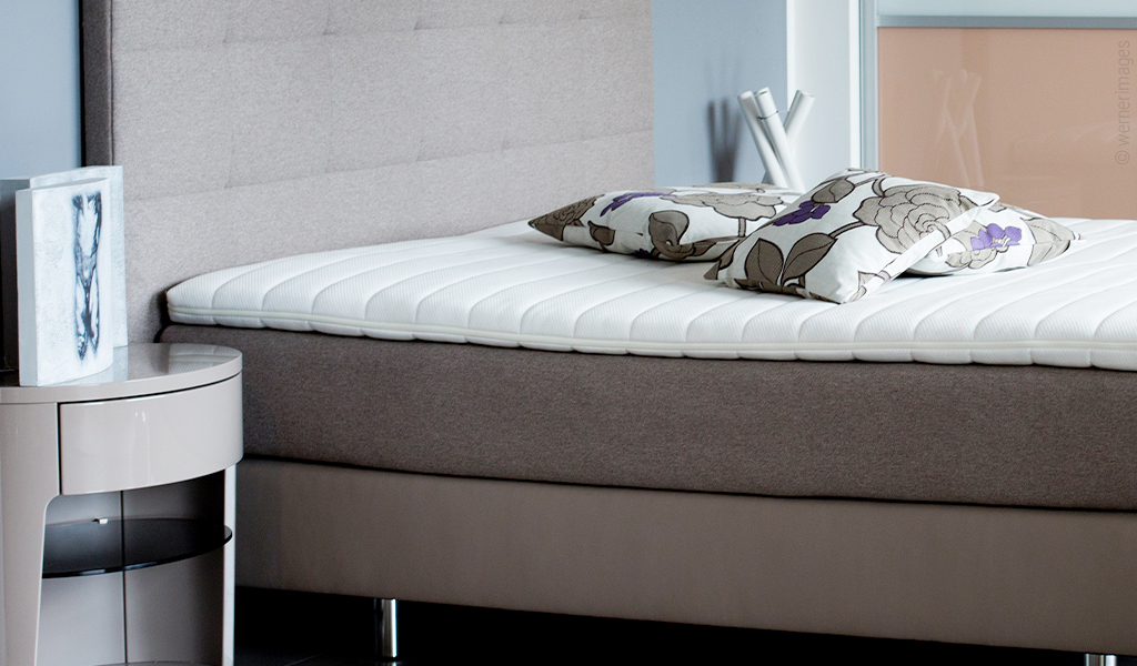 Auf einem Polsterbett liegt eine Boxspringmatratze. Darauf liegen ein Topper und drei dekorative Kissen.